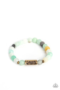 Meeting at the center of an elastic stretch bracelet, a brass, rectangular bar, hammered in an antiqued sheen, is stamped with the word "peace" for a serene statement. Brass accents, and milky stone beads, in various shades of blue, white, gray, and yellow, encircle the brass bar creating a calming, cheerful finish. As the stone elements in this piece are natural, some color variation is nor